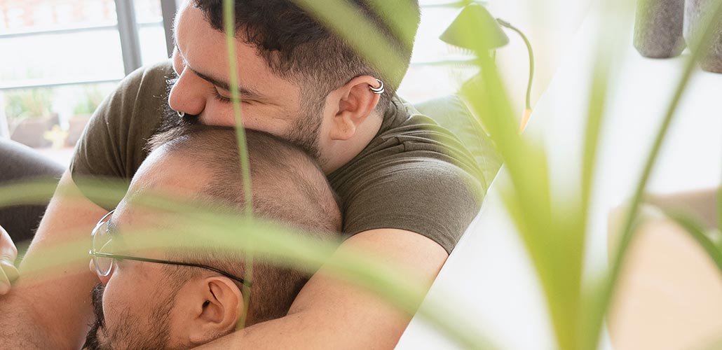 two men cuddling on couch with houseplant out of focus in foreground