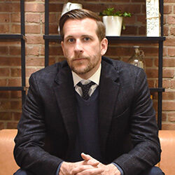 A photo of Kevin Vail wearing a suit, sitting on a couch in front of a brick wall and shelving