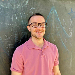 A photo of Dr. Andrew Bingman smiling in front of a slate wall with drawings on it