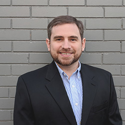 A photo of Jason Stotts wearing a black blazer and collared shirt, in front of a white brick wall