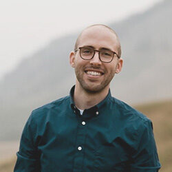 A photo of Joel Mader smiling in front of a blurry mountain backdrop