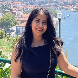 A photo of Maheema Dua smiling while resting against a handrail, with a scenic view of water behind